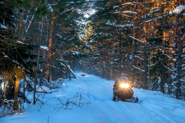 Two snowmobiles in single file along a snowy trail in Nova Scotia. The sun shines through the trees as the snowmobiles pass. 